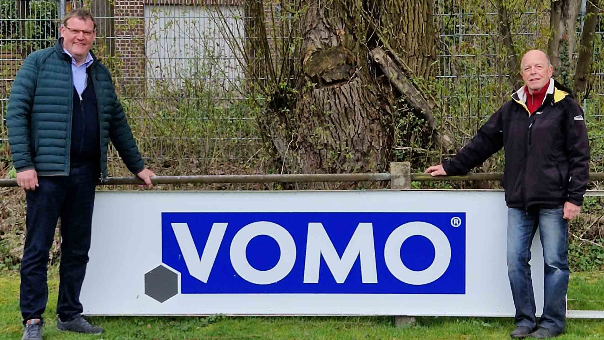 Zwei Männer stehen links und rechts neben einem großen Schild mit dem blauen VOMO-Logo. Im Hintergrund befinden sich Bäume und ein Metallzaun.