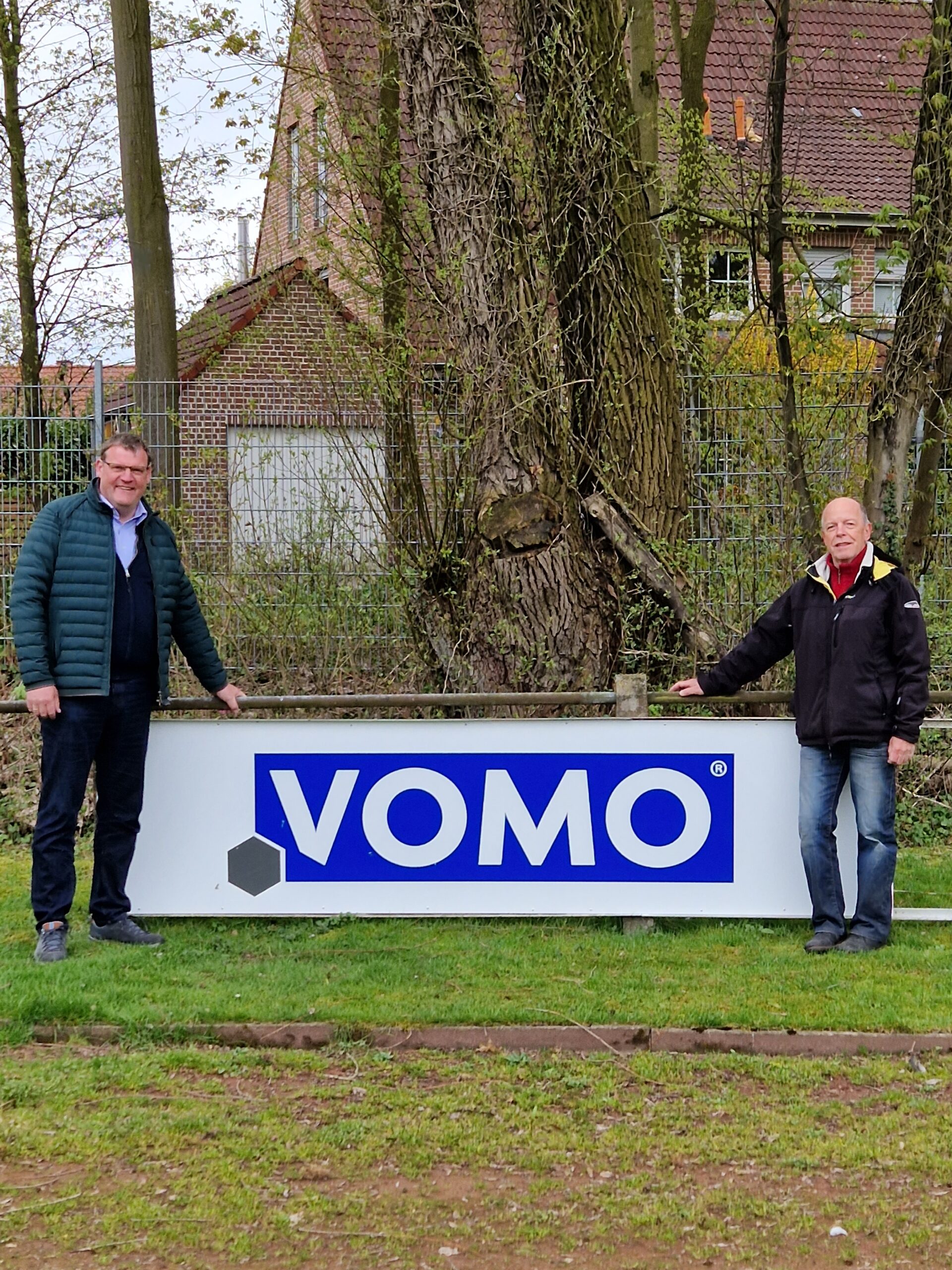 Zwei Männer stehen im Freien neben einem großen Schild mit dem Firmenlogo ‚VOMO‘, das blau-weiß gestaltet ist und einen hexagonalen Akzent enthält. Im Hintergrund sind hohe Bäume sowie Backsteingebäude sichtbar, was auf eine ländliche oder suburbane Umgebung hindeutet. Beide Männer tragen legere Kleidung und lächeln freundlich in die Kamera.